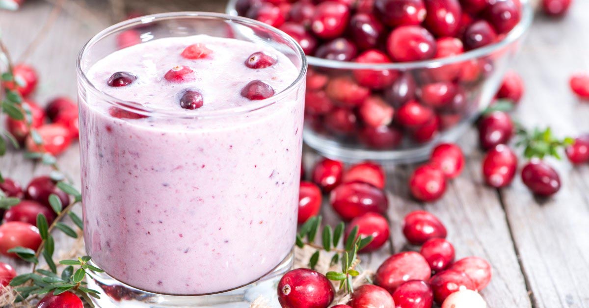 A cranberry smoothie in a glass with cranberries floating on top. The glass is surrounded by cranberries on a wooden table and by a clear glass bowl of cranberries.