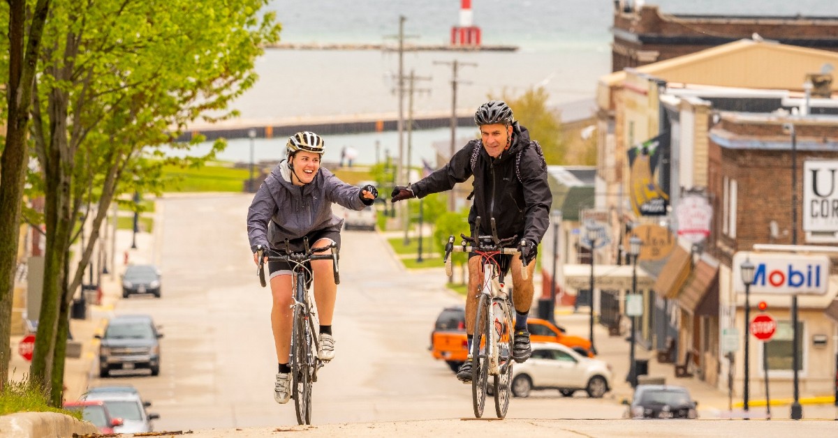 Woman and man on bicycles bump fists while riding up a steep hill in a small town.